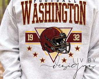 May include: A light grey sweatshirt with a burgundy and gold football helmet design. The word "WASHINGTON" is above the helmet, with "FOOTBALL" above it. The numbers "19" and "32" are on either side of the helmet.