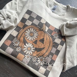 May include: Cream-coloured sweatshirt with a pumpkin design, daisy flowers, and a chequered background. The pumpkin has a carved face. The design is printed on the front. The sleeves are rolled up.