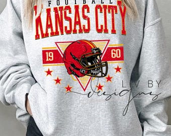 Peut inclure: Un sweat-shirt gris avec l'inscription "FOOTBALL KANSAS CITY" en rouge et jaune. Un casque de football rouge est centré en dessous, avec les chiffres "19" et "60" de chaque côté. Plusieurs étoiles rouges sont dispersées autour du casque.