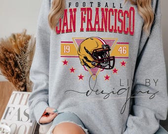 Puede incluir: Una sudadera gris con un diseño de fútbol. El diseño incluye las palabras "FOOTBALL SAN FRANCISCO" en rojo y dorado, un casco de fútbol, los números "19" y "46" y estrellas rojas. La sudadera es de cuello redondo y manga larga.