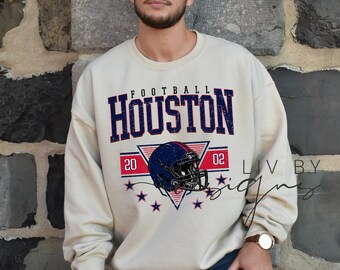 Könnte beinhalten: Cremefarbenes Sweatshirt mit dem Wort "HOUSTON" in Marineblau, über einer Footballhelm-Grafik. Die Grafik enthält die Zahlen "20" und "02" und mehrere blaue Sterne. Das Sweatshirt ist ein lässiger Stil.