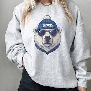 May include: A light gray sweatshirt featuring a polar bear wearing a blue beanie with the word "TORONTO" and sunglasses. The bear has a blue scarf. The sweatshirt is a casual style.