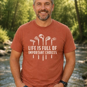 Puede incluir: Una camiseta de color &oacute;xido con el texto blanco "LIFE IS FULL OF IMPORTANT CHOICES", junto con gr&aacute;ficos de palos de golf. La camiseta la lleva una persona con barba gris y pelo oscuro.