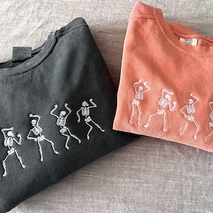Puede incluir: Dos sudaderas con esqueletos bordados bailando. Una sudadera es negra y la otra es de color naranja claro.