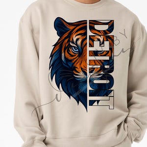 May include: Beige sweatshirt with a tiger graphic in orange and blue. The word "DETROIT" is printed vertically in a bold font. The design is centered on the front of the sweatshirt, offering a striking visual.