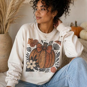 May include: Cream-colored sweatshirt featuring a large orange pumpkin graphic with a bow, surrounded by floral and leaf elements. The design has a rustic, fall-themed aesthetic. The long-sleeved top is made of a soft material.