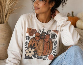 May include: Cream-colored sweatshirt featuring a large orange pumpkin graphic with a bow, surrounded by floral and leaf elements. The design has a rustic, fall-themed aesthetic. The long-sleeved top is made of a soft material.
