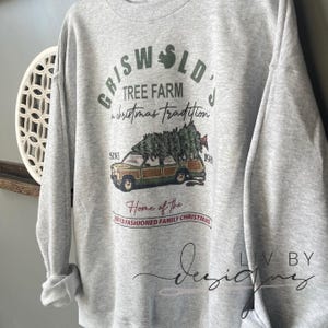 May include: A gray sweatshirt with a Christmas-themed design featuring a station wagon with a Christmas tree on top. The text on the sweatshirt reads "Griswold's Tree Farm a Christmas Tradition Since 198 Home of the Old Fashioned Family Christmas".