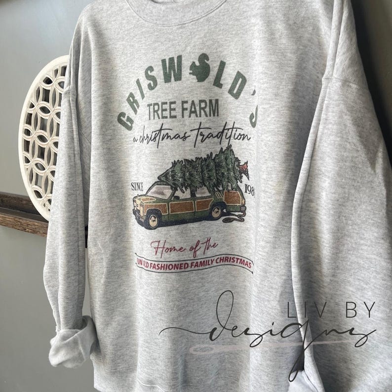 May include: A gray sweatshirt with a Christmas-themed design featuring a station wagon with a Christmas tree on top. The text on the sweatshirt reads "Griswold's Tree Farm a Christmas Tradition Since 198 Home of the Old Fashioned Family Christmas".