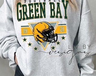 May include: A grey sweatshirt featuring the text "FOOTBALL GREEN BAY" in green and gold. A yellow and black football helmet is below the text, with the years "19" and "21" on either side. The design includes stars.