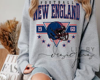 Könnte beinhalten: Graues Sweatshirt mit einem verblassten Aufdruck "FOOTBALL NEW ENGLAND 1960". Das Design umfasst einen blauen Footballhelm, rote und blaue Streifen und blaue Sterne. Das Sweatshirt hat einen Rundhalsausschnitt und lange Ärmel.