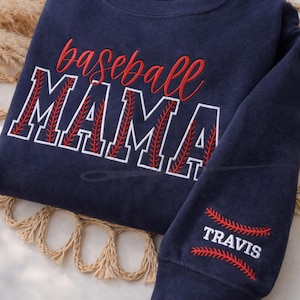 May include: Navy blue sweatshirt with the words "baseball MAMA" embroidered in red and white. The sleeve features the name "TRAVIS" with baseball stitching. A casual top for baseball fans.