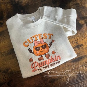 Könnte beinhalten: Ein hellgraues Sweatshirt mit dem Text "Cutest Pumpkin in the Patch" und einem Cartoon-Kürbis mit Sonnenbrille und Schleife. Der Kürbis ist orange mit braunen Schuhen und Armen. Das Sweatshirt liegt auf einer Holzoberfläche.
