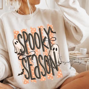 May include: A cream-colored crewneck sweatshirt featuring a graphic print with the text "SPOOKY SEASON" in black script. The design includes two white ghosts, small black bats, and vertical orange stripes with white polka dots as a background.