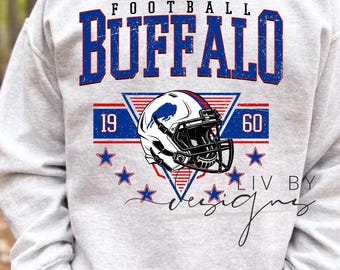 May include: A light grey sweatshirt with a graphic design featuring the word "FOOTBALL BUFFALO" in blue, a football helmet, and the numbers "1960". The design includes red and blue accents and stars.