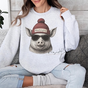 May include: A light gray sweatshirt featuring a pig wearing a maroon beanie with "Washington" on it and black sunglasses. The pig is smiling. The sweatshirt has the words "Liv by Design" in cursive.
