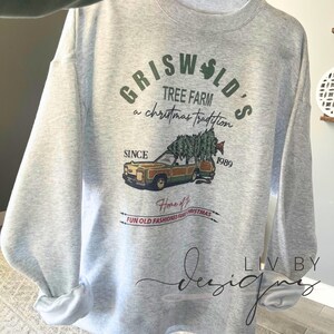 May include: A gray sweatshirt with a Christmas-themed design featuring a station wagon with a Christmas tree on top. The text on the sweatshirt reads "Griswold's Tree Farm a Christmas Tradition Since 1989 Home of Fun Old Fashioned Family Christmas".