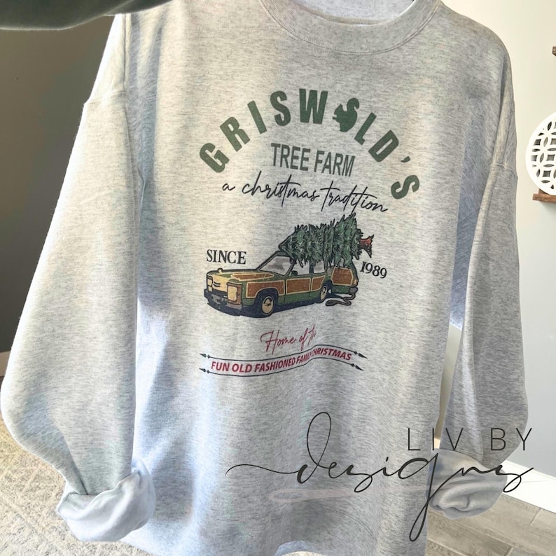 May include: A gray sweatshirt with a Christmas-themed design featuring a station wagon with a Christmas tree on top. The text on the sweatshirt reads "Griswold's Tree Farm a Christmas Tradition Since 1989 Home of Fun Old Fashioned Family Christmas".