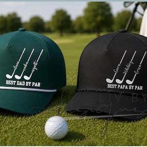 May include: Two golf caps, one dark green and one black, with white text and golf club graphics. The green cap says "BEST DAD BY PAR" with names. The black cap says "BEST PAPA BY PAR" with names. A golf ball rests on the grass.