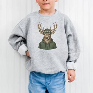 May include: A light gray sweatshirt featuring a deer wearing a green hoodie and a green hat with the word "Packers". The deer has large antlers and a brown face. The sweatshirt has white cuffs and is worn by a child.