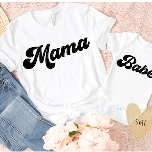 Puede incluir: Camiseta blanca con la palabra "Mama" en fuente retro negra, combinada con un body blanco para bebé con "Babe" en la misma fuente. Los artículos se muestran con vaqueros azules, flores y una etiqueta en forma de corazón.