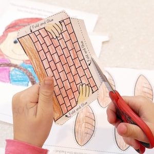 Pu&ograve; includere: Una mano di bambino tiene un'attivit&agrave; manuale di carta di un muro di mattoni con istruzioni per piegare e incollare. L'attivit&agrave; manuale di carta viene ritagliata con delle forbici rosse. L'attivit&agrave; manuale di carta fa parte di un progetto pi&ugrave; ampio che include pagnotte di pane.