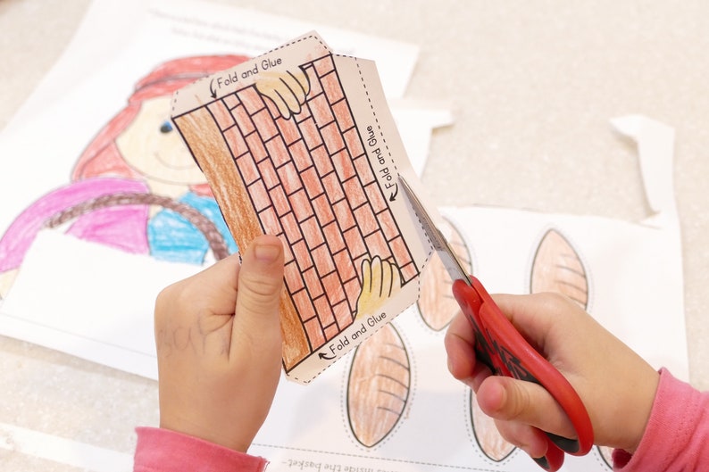 Pu&ograve; includere: Una mano di bambino tiene un'attivit&agrave; manuale di carta di un muro di mattoni con istruzioni per piegare e incollare. L'attivit&agrave; manuale di carta viene ritagliata con delle forbici rosse. L'attivit&agrave; manuale di carta fa parte di un progetto pi&ugrave; ampio che include pagnotte di pane.