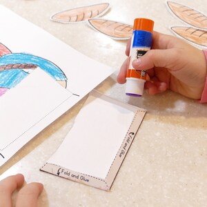 Pu&ograve; includere: Una mano di bambino tiene un bastoncino di colla sopra un'attivit&agrave; di carta bianca con le parole "Piega e incolla" e "Piega pi&ugrave; incolla" stampate su di essa. L'attivit&agrave; &egrave; accanto a un disegno di una ragazza con i capelli rossi e una camicia blu.