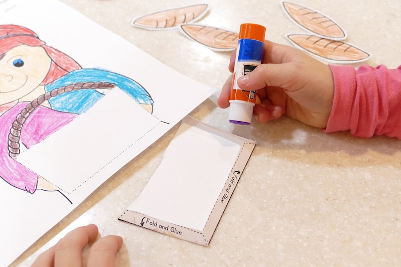 Pu&ograve; includere: Una mano di bambino tiene un bastoncino di colla sopra un'attivit&agrave; di carta bianca con le parole "Piega e incolla" e "Piega pi&ugrave; incolla" stampate su di essa. L'attivit&agrave; &egrave; accanto a un disegno di una ragazza con i capelli rossi e una camicia blu.