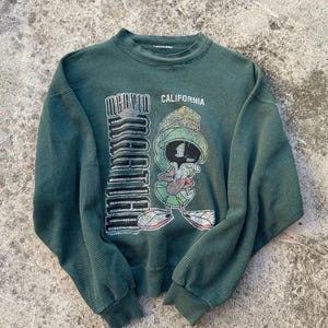 May include: Green ribbed crew neck sweatshirt featuring a graphic of Marvin the Martian with the text "MARVIN CALIFORNIA". The sweatshirt has long sleeves and a relaxed fit. The graphic includes the character's face, helmet, and a number 4.
