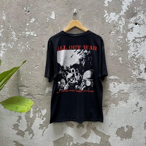 Puede incluir: Camiseta negra con la frase "ALL OUT WAR" en rojo sobre un gráfico en blanco y negro de figuras con máscaras de gas. El texto "FOR THOSE WHO WERE CRUCIFIED" está debajo.