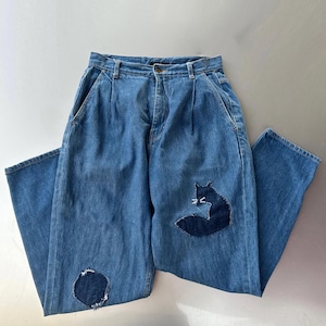 Może przedstawiać: Niebieskie dżinsy z wysokim stanem i aplikacją lisa na prawej nogawce. Dżinsy mają łatkę na lewym kolanie. Dżinsy są złożone na białej powierzchni.