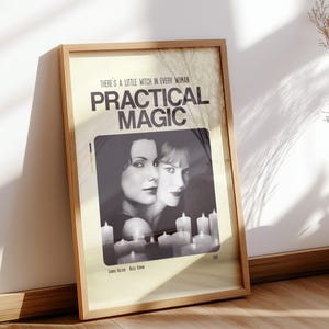 Peut inclure: Une affiche encadrée avec le texte "PRACTICAL MAGIC" et la phrase "THERE IS A LITTLE WITCH IN EVERY WOMAN." L'affiche présente une image en noir et blanc de deux femmes et des bougies. Un vase avec des plantes séchées est posé à côté de l'affiche.