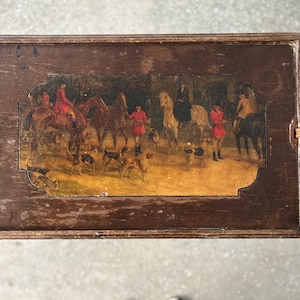 May include: A rectangular wooden serving tray with a painted hunting scene. The artwork depicts people on horseback, dogs, and figures in red coats. The tray has dark brown wooden handles and a distressed finish.