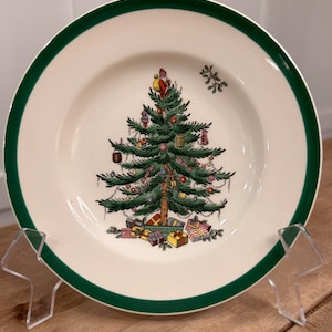 May include: A white ceramic Christmas plate with a green rim features a colorful illustration of a Christmas tree decorated with ornaments, presents, and a small figure at the top.
