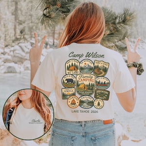 May include: A cream-colored t-shirt featuring a large graphic on the back with the text Camp Wilson and Lake Tahoe 2026, surrounded by various camping-themed badges. The front chest area displays the text Charlie Mosquito Magnet.
