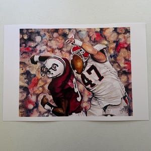 May include: A watercolor-style art print depicting two American football players in action. One player wears a maroon jersey and white helmet, while the other wears a white jersey with the number 47 and a red helmet, reaching for a football.