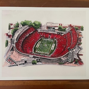 May include: Watercolor print of a football stadium with red seating and a green field. The stadium has the word "GEORGIA" on the field and the text "SATURDAY IN ATHENS" on a sign. The print is on white paper.
