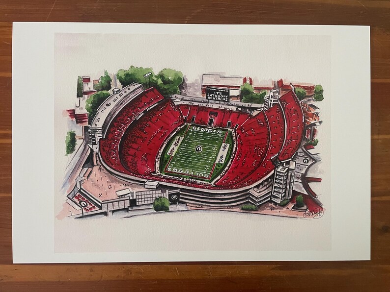 May include: Watercolor print of a football stadium with red seating and a green field. The stadium has the word "GEORGIA" on the field and the text "SATURDAY IN ATHENS" on a sign. The print is on white paper.