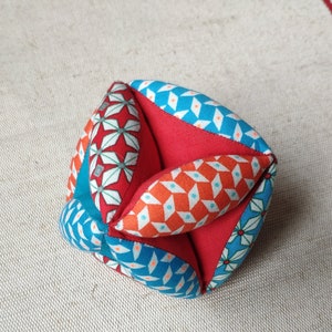 May include: A colorful fabric cube with a geometric pattern in shades of red, blue, and white. The cube is made of multiple fabric panels sewn together.