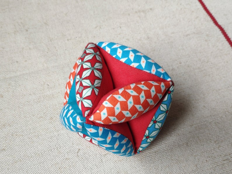 May include: A colorful fabric cube with a geometric pattern in shades of red, blue, and white. The cube is made of multiple fabric panels sewn together.