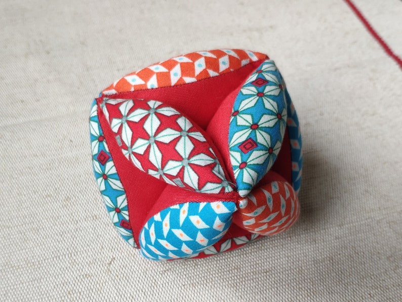 May include: A red, blue, and orange fabric cube with a geometric pattern. The cube is made of multiple fabric panels sewn together.