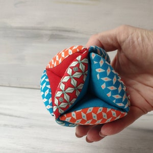 May include: A colorful fabric ball with a geometric pattern in shades of red, blue, and orange. The ball is made of multiple fabric panels sewn together.