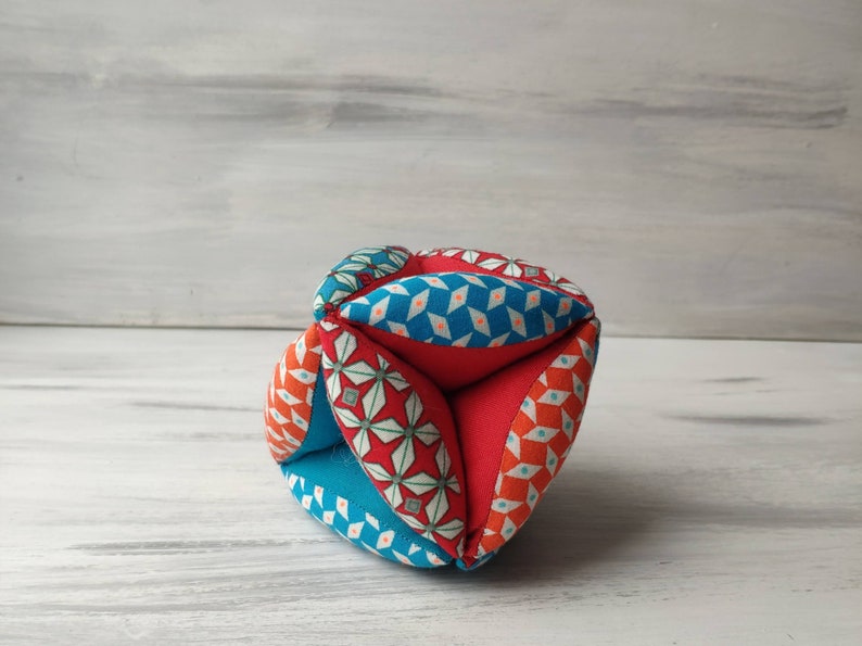 May include: A multi-colored fabric ball with a geometric pattern. The ball is made of red, blue, and white fabric and has a diamond pattern on one side and a floral pattern on the other.