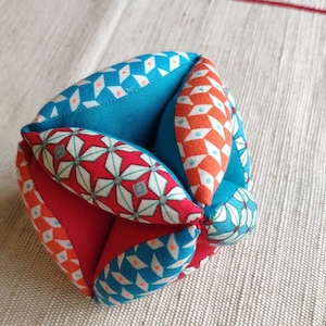 May include: A colorful fabric ball with a geometric pattern in shades of red, blue, and orange. The ball is made of multiple fabric pieces sewn together.