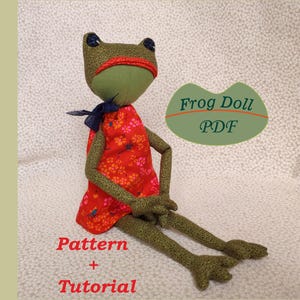 May include: A fabric frog doll with olive green speckled skin, wearing a red dress with a pink and orange floral pattern. The doll has a dark blue bow at the neck and a red headband. Text on the image reads Frog Doll PDF, Pattern + Tutorial.