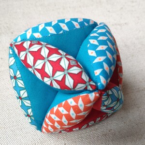 May include: A turquoise blue fabric cube with red, white, and orange geometric patterns. The cube is made of multiple fabric panels sewn together.