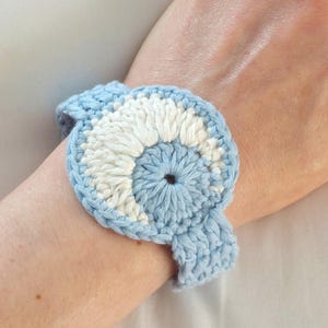 Könnte beinhalten: Ein gehäkeltes Armband in Hellblau und Weiß mit einem kreisförmigen Design. Das Armband ist handgefertigt mit einem strukturierten Stich und wird am Handgelenk getragen. Die Farbpalette ist weich und das Design ist aufwendig, was auf einen Bastelartikel hindeutet.