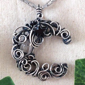 May include: A silver-coloured crescent moon pendant necklace. The pendant has a wire-wrapped design with a clear, round stone and a small star. The necklace is on a silver chain, and the background is a light beige fabric.