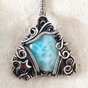 May include: A silver pendant necklace. The triangular pendant has a light blue stone, encased in a wire-wrapped design with decorative swirls and a small star. The chain is silver. The pendant is approximately 3 cm in height.
